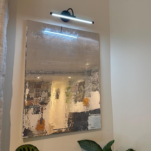 May include: A rectangular wall mirror covered in small square silver mirrored tiles, reflecting a room interior. Above the mirror, a black horizontal LED wall light fixture is mounted, casting a bright white glow downwards onto the surface.