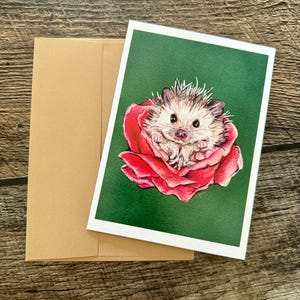 May include: A greeting card featuring a detailed illustration of a hedgehog nestled within a red rose. The card has a white border and a green background. A brown envelope is included.