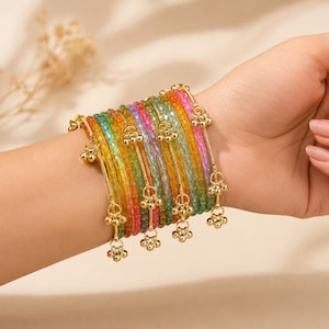 May include: A selection of vibrant beaded bracelets in shades of yellow, green, pink, and blue are displayed on a wrist. Each bracelet is adorned with a gold-coloured charm featuring a floral design. The bracelets are a colourful accessory.