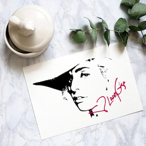 May include: Black and white illustration of a woman wearing a hat with a pink signature that says "Lady Gaga".