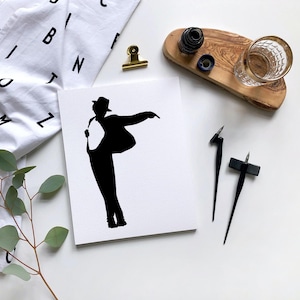 May include: A black silhouette of a person in a hat and jacket, striking a pose on a white card. The image is surrounded by calligraphy tools, a glass, and a wooden tray. A white cloth with black letters is in the background.