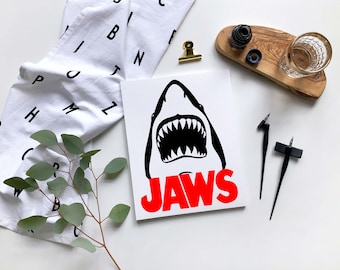 May include: A white canvas print with a black silhouette of a shark's open mouth with sharp teeth. The text "JAWS" is in red letters below the shark.