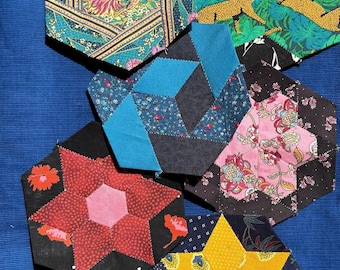 May include: A collection of six hexagonal fabric pieces with various patterns. Designs include floral, geometric, and animal prints in colors like teal, pink, yellow, and black. The pieces are arranged on a blue surface.