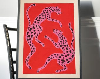 May include: A framed art print featuring two pink leopards with black spots positioned against a solid red background. The leopards are depicted in dynamic, leaping poses. The artwork is enclosed in a simple, thin black frame with a cream-colored mat.
