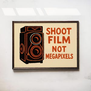 May include: Framed art print featuring a vintage camera illustration in black and orange on a cream background. The text reads "SHOOT FILM NOT MEGAPIXELS". The artwork is encased in a dark brown frame.