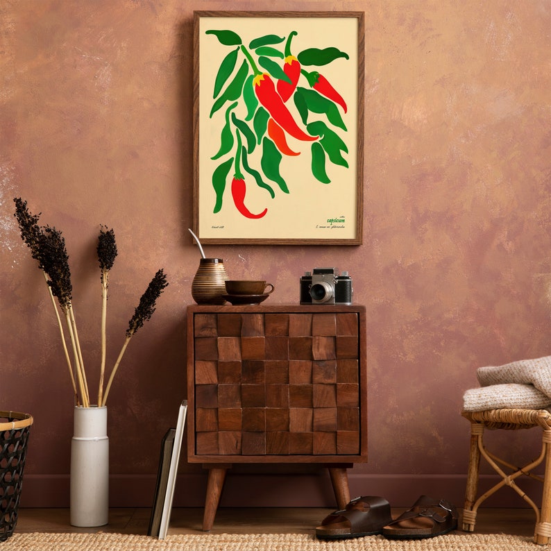 Op de afbeelding: Een ingelijst afdruk van een rode chilipeperplant met groene bladeren op een cr&egrave;mekleurige achtergrond. De afdruk hangt aan een muur met een bruin houten kastje en een rieten stoel op de voorgrond.