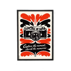May include: Framed art print featuring a black and white camera illustration with red and black decorative elements. The text "Capture the moment, cherish the memories" is displayed in a stylized font.