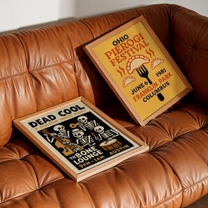 K&ouml;nnte beinhalten: Zwei gerahmte Poster mit Holzrahmen liegen auf einem braunen Ledersofa. Ein Poster ist gelb mit dem Text "OHIO PIEROGI FESTIVAL" und "JUNE 6 1981 FRANKLIN PARK COLUMBUS". Das andere ist schwarz mit einer Skelettband und dem Text "DEAD COOL A SKELETON SWING NIGHT."