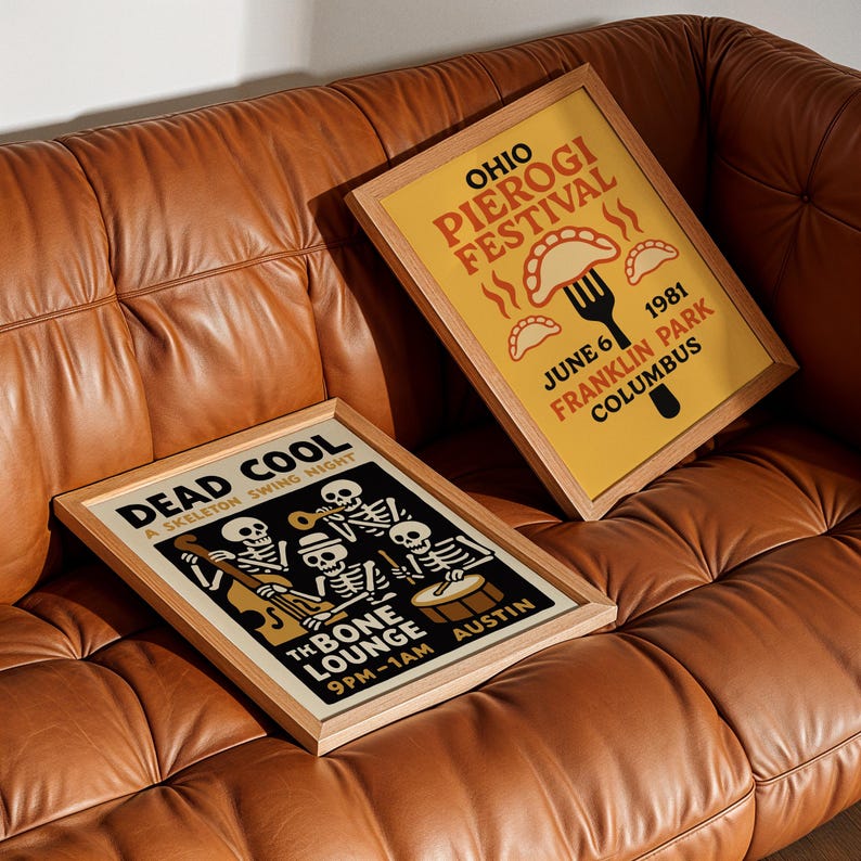 K&ouml;nnte beinhalten: Zwei gerahmte Poster mit Holzrahmen liegen auf einem braunen Ledersofa. Ein Poster ist gelb mit dem Text "OHIO PIEROGI FESTIVAL" und "JUNE 6 1981 FRANKLIN PARK COLUMBUS". Das andere ist schwarz mit einer Skelettband und dem Text "DEAD COOL A SKELETON SWING NIGHT."