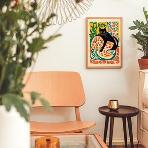 May include: A framed art print featuring a black cat lounging on a pizza, with a colorful background of flowers and abstract shapes. The artwork is displayed on a white wall in a room with a wooden chair and side table.