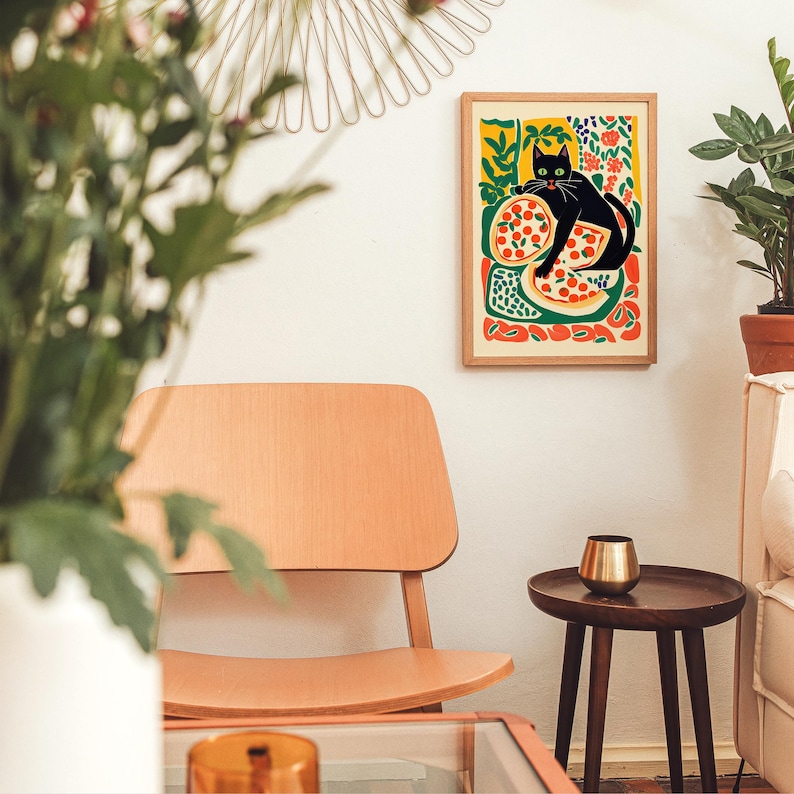 May include: A framed art print featuring a black cat lounging on a pizza, with a colorful background of flowers and abstract shapes. The artwork is displayed on a white wall in a room with a wooden chair and side table.