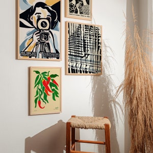 Op de afbeelding: Vier ingelijste prints op een witte muur. De print linksboven toont een vrouw in een geel shirt die een camera vasthoudt. De print rechtsboven toont een zwart-witte illustratie van een stadsbeeld. De print linksonder toont een rode en groene illustratie van chilipepers. De print rechtsonder is leeg.