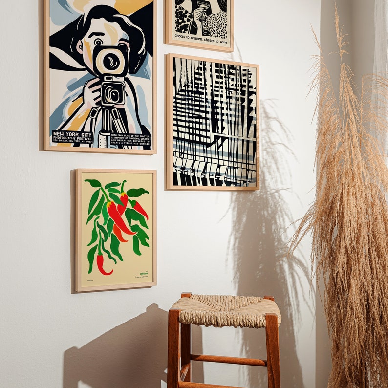 Op de afbeelding: Vier ingelijste prints op een witte muur. De print linksboven toont een vrouw in een geel shirt die een camera vasthoudt. De print rechtsboven toont een zwart-witte illustratie van een stadsbeeld. De print linksonder toont een rode en groene illustratie van chilipepers. De print rechtsonder is leeg.