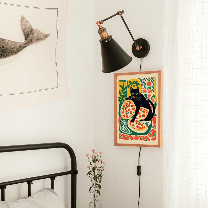 May include: A framed art print featuring a black cat with green eyes lounging on a pizza, set against a colorful floral background. The print is mounted on a light wood frame and hangs on a white wall. A black wall lamp is mounted above the print.