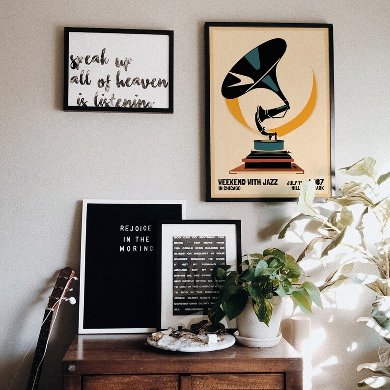 May include: A black and white framed sign with the text "Rejoice in the Morning" on a wooden shelf with a guitar, a white framed sign with the text "Speak up all of heaven is listening", a black framed poster with a vintage illustration of a gramophone and the text "Weekend with Jazz in Chicago" and a potted plant.