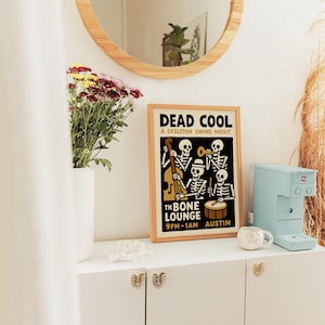 K&ouml;nnte beinhalten: Ein gerahmtes Poster mit dem Text "DEAD COOL" und "A SKELETON SWING NIGHT" zeigt eine Band von Skelettmusikern. Eine hellblaue Kaffeemaschine und eine kleine wei&szlig;e Tasse stehen auf einem wei&szlig;en Schrank. Eine Vase mit Blumen und ein runder Spiegel sind ebenfalls zu sehen.