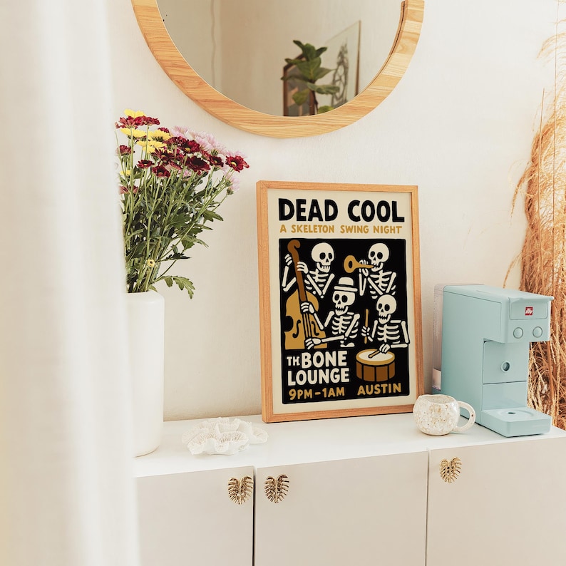 K&ouml;nnte beinhalten: Ein gerahmtes Poster mit dem Text "DEAD COOL" und "A SKELETON SWING NIGHT" zeigt eine Band von Skelettmusikern. Eine hellblaue Kaffeemaschine und eine kleine wei&szlig;e Tasse stehen auf einem wei&szlig;en Schrank. Eine Vase mit Blumen und ein runder Spiegel sind ebenfalls zu sehen.