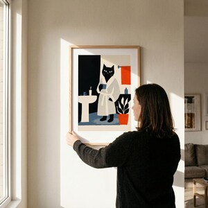 Puede incluir: Una impresi&oacute;n art&iacute;stica enmarcada representa un gato negro con una bata blanca sosteniendo una taza en un ba&ntilde;o. La obra de arte presenta un estilo minimalista con una paleta de colores negro, blanco, naranja y azul. El gato est&aacute; cerca de un lavabo y una planta en maceta.