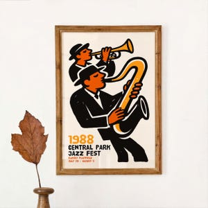 May include: A framed poster for the 1988 Central Park Jazz Fest. The poster features two musicians in black suits and hats, one playing a trumpet and the other a saxophone. The poster has a cream background with orange and black graphics.