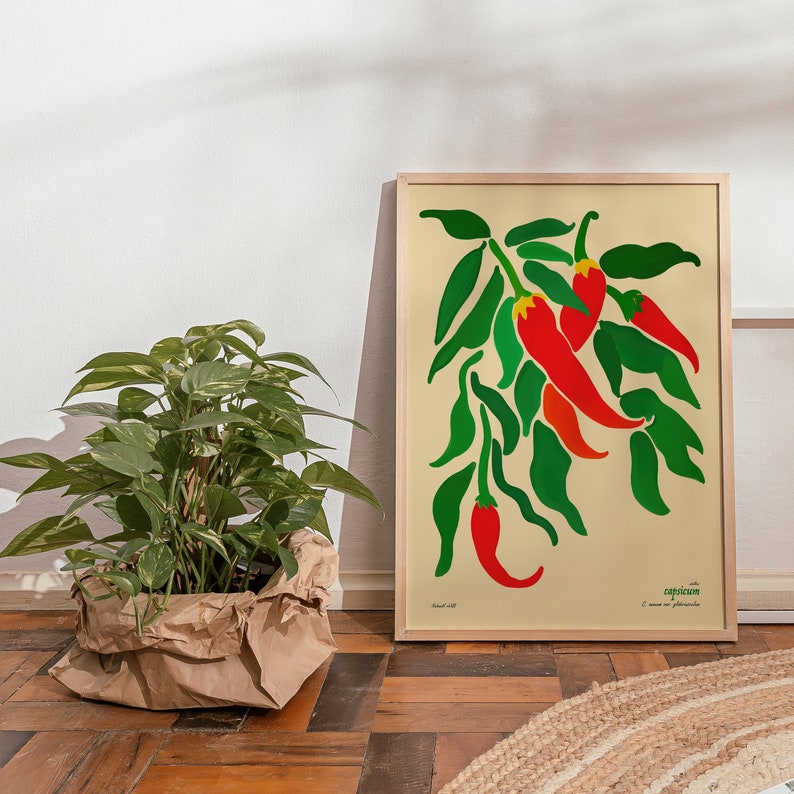 Op de afbeelding: Een ingelijste print van een gestileerde illustratie van rode pepers met groene bladeren. De print is getiteld "Capsicum" en heeft de tekst "C. annuum var. glabriusculum".