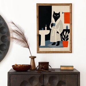 Puede incluir: Una impresi&oacute;n art&iacute;stica enmarcada representa un gato negro con una bata blanca sosteniendo una taza en un ba&ntilde;o. La obra de arte presenta un dise&ntilde;o minimalista con una paleta de colores negro, blanco, azul y naranja. La impresi&oacute;n se muestra encima de un mueble de madera oscura.
