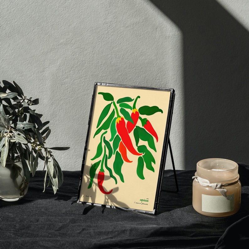 Op de afbeelding: Een ingelijste print van een gestileerde illustratie van rode pepers met groene bladeren op een cr&egrave;mekleurige achtergrond. De print is in een zwart frame en heeft de tekst "Capsicum" onderaan.