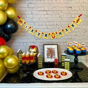 Könnte beinhalten: Ein Partytisch, der mit einem Iron-Man-Thema dekoriert ist. Der Tisch hat eine schwarz-goldene Tischdecke, ein rot-goldenes Banner mit der Aufschrift "Iron Man" und eine Vielzahl von Iron-Man-Dekorationen, darunter Cupcakes, Kekse und eine kleine Iron-Man-Figur.