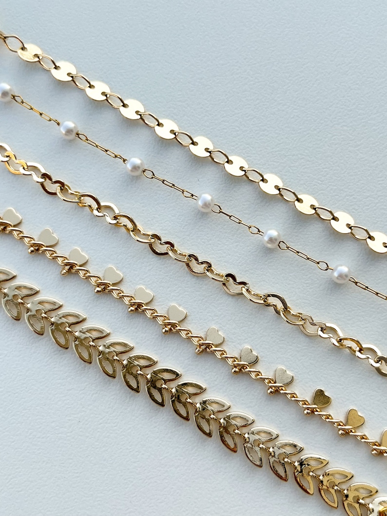 May include: Five gold chains with different styles, including a chain with small round links, a chain with small heart charms, a chain with small leaf charms, and a chain with small pearl accents.