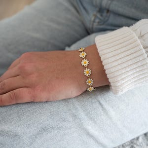 Może przedstawiać: Złota bransoletka z łańcuszkiem i białymi zawieszkami w kształcie stokrotek. Bransoletka jest na nadgarstku osoby.
