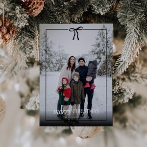 Pode incluir: Cart&atilde;o de Natal com uma foto de fam&iacute;lia numa floresta nevada. O cart&atilde;o tem uma borda preta com um la&ccedil;o e a inscri&ccedil;&atilde;o "merry CHRISTMAS". A fam&iacute;lia est&aacute; vestida com roupas de inverno.