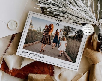 Könnte beinhalten: Eine rechteckige Weihnachtsgrußkarte mit einem Foto einer Familie auf einem Holzsteg. Die Karte zeigt eine schwarze Schleifengrafik, den Text merry CHRISTMAS und THE WILLIAMSON'S | 202X. Ein weißer Umschlag und ein Band sind zu sehen.