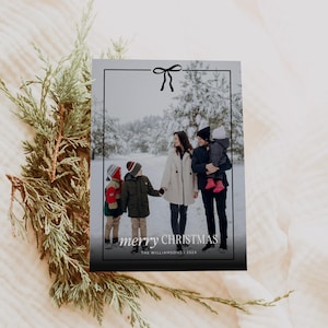 Pode incluir: Um cart&atilde;o de Natal com uma foto de fam&iacute;lia numa floresta nevada. O cart&atilde;o tem uma borda preta com um la&ccedil;o e as palavras "merry CHRISTMAS" em branco. A fam&iacute;lia est&aacute; vestida com roupas de inverno.