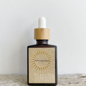 K&ouml;nnte beinhalten: Eine braune Glasflasche mit einem Bambusdeckel und einer wei&szlig;en Tropfpipette. Die Flasche hat ein Etikett mit einer Sonnengrafik und dem Text "ashwagandha" und "STARBORN".