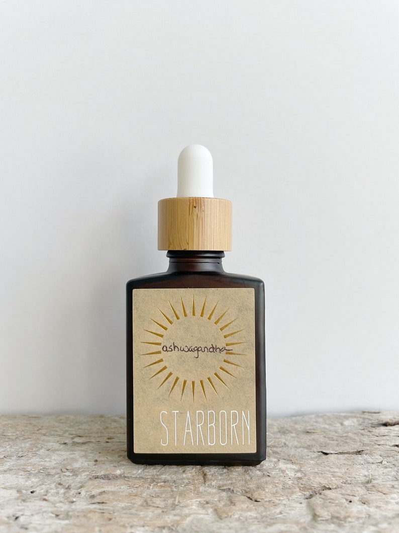 K&ouml;nnte beinhalten: Eine braune Glasflasche mit einem Bambusdeckel und einer wei&szlig;en Tropfpipette. Die Flasche hat ein Etikett mit einer Sonnengrafik und dem Text "ashwagandha" und "STARBORN".
