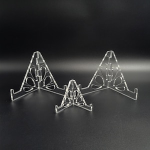 Op de afbeelding: Drie heldere acryl display stands van verschillende maten. Elke standaard heeft een driehoekige vorm met decoratieve uitsparingen en een kleine richel om items vast te houden. De stands zijn gerangschikt op een zwart oppervlak.