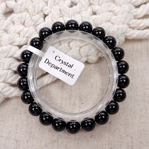 May include: A circular bracelet made of polished black spherical beads strung together. A white rectangular tag is attached to the bracelet, featuring the printed text "Crystal Department" with a small star symbol above the word Crystal.