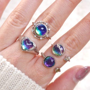 May include: A collection of silver rings with iridescent, color-shifting stones. The rings feature various designs, including a heart, a round setting with a star, and a round setting with a decorative border. The stones display a range of colors, including blue, purple, and green.