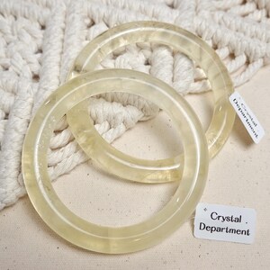 May include: Two clear yellow crystal bracelets, one slightly larger than the other, on a white crocheted surface. The bracelets are thick and round, with a smooth, polished finish. A white tag with black text reads "Crystal Department".