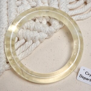 May include: A clear, translucent bangle bracelet made of glass or plastic. The bangle is thick and has a smooth, polished finish.