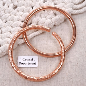 May include: Two copper-colored metal bracelets. One is a smooth, closed circular bangle, and the other is an open-ended cuff with a hammered, textured surface. A small white rectangular tag with the text Crystal Department is visible.