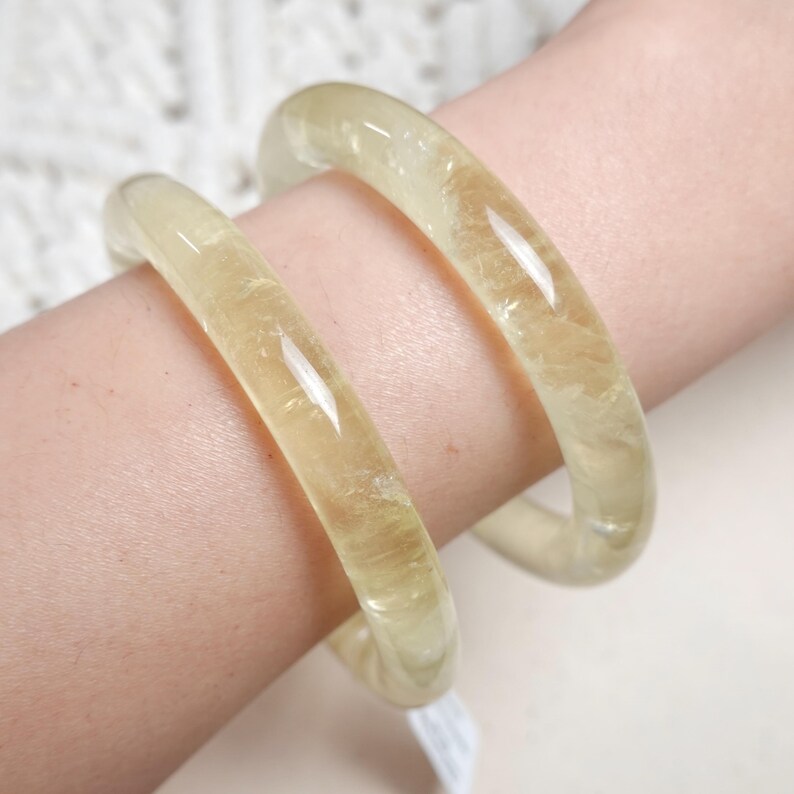 May include: Two yellow citrine crystal bangles, one slightly larger than the other, worn on a person's wrist.