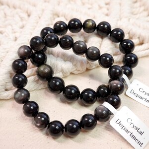 May include: Two black gemstone bracelets with gold flecks. The bracelets are made of round beads and are stacked on top of each other. The bracelets are on a white crocheted surface with a white tag that says "Crystal Department".