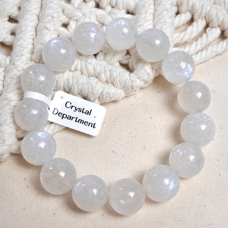 May include: White moonstone beaded bracelet with iridescent shimmer. The bracelet is made of round beads and is approximately 8 inches in length. The bracelet is on a white background with a tag that reads "Crystal Department."