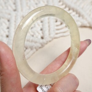 May include: A translucent, pale yellow bangle bracelet made of natural selenite crystal. The bangle is smooth and polished, with a slightly oval shape.