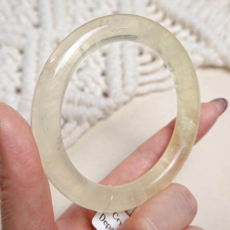 May include: A translucent, pale yellow bangle bracelet made of natural selenite crystal. The bangle is smooth and polished, with a slightly oval shape.