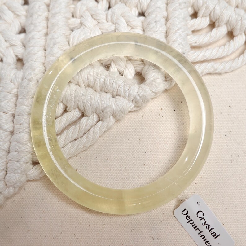 May include: A pale yellow, translucent bangle bracelet made of natural crystal. The bracelet is lying on a white crocheted surface. The text on the tag reads "Crystal Department."