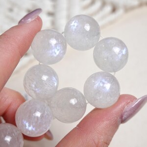 May include: A strand of white iridescent moonstone beads. The beads are round and have a smooth, polished finish. The beads are strung on a clear elastic cord.