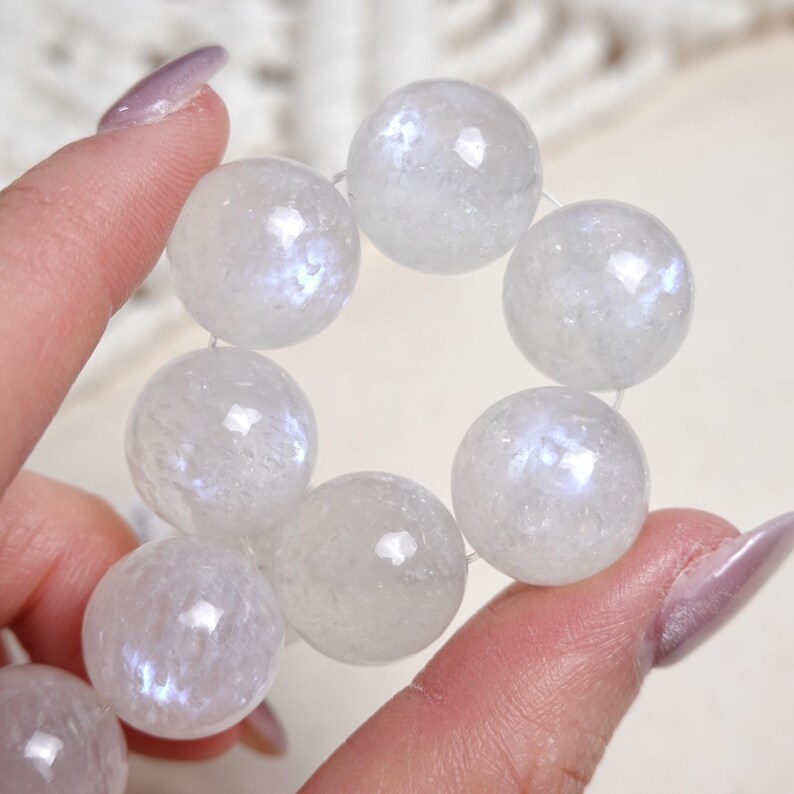 May include: A strand of white iridescent moonstone beads. The beads are round and have a smooth, polished finish. The beads are strung on a clear elastic cord.