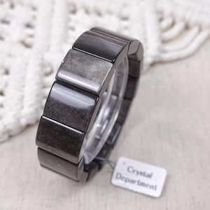May include: A bracelet composed of rectangular, polished dark grey obsidian stones with a subtle metallic sheen. The bracelet features a small white rectangular tag attached by a thin string, clearly printed with the text Crystal Department.