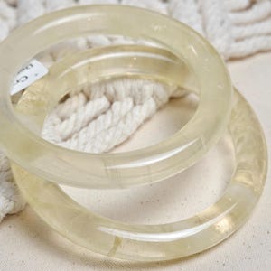 May include: Two clear, translucent bangle bracelets made of polished, light yellow stone. The bracelets are stacked on top of each other.